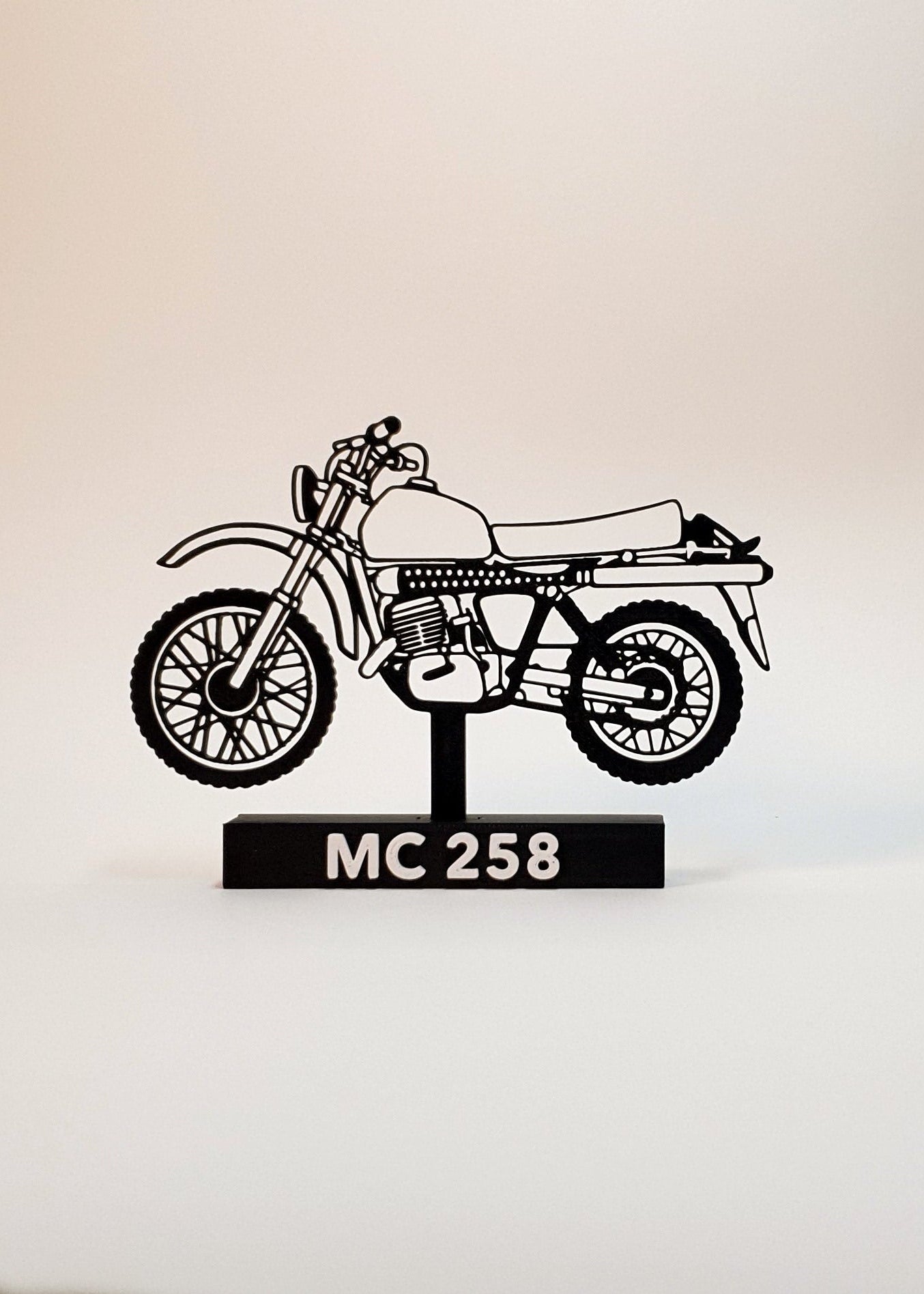 A black metal cutout of a classic motorcycle on a stand with the text 'MC 258' on a black base, set against a plain light beige background.