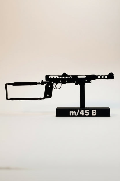 A black metal silhouette model of an m/45 B machine gun mounted on a black display base with white text, set against a plain off-white background.