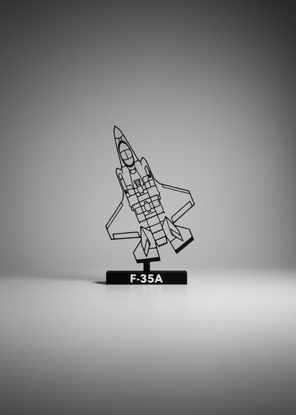 A black plastic F-35A jet model on a black stand, displayed on a white table against a gray wall with a framed picture in the background.