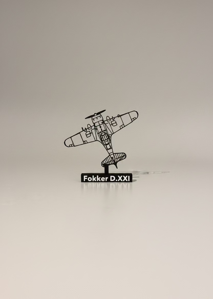 A black plastic sculpture of a Fokker D.XXI airplane displayed on a white table against a gray wall, with a framed picture in the background.