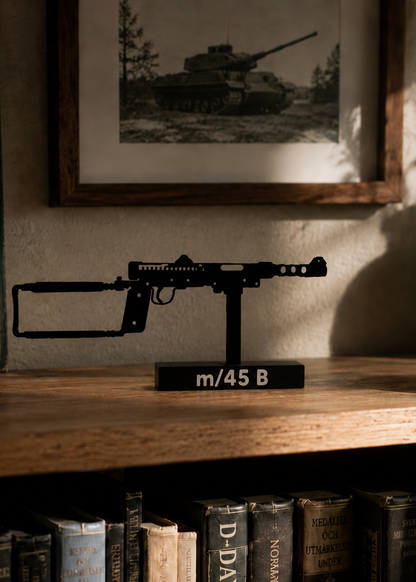 A black metal model of an m/45 Browning machine gun mounted on a black display stand, placed on a wooden shelf in front of a framed black-and-white military photograph.